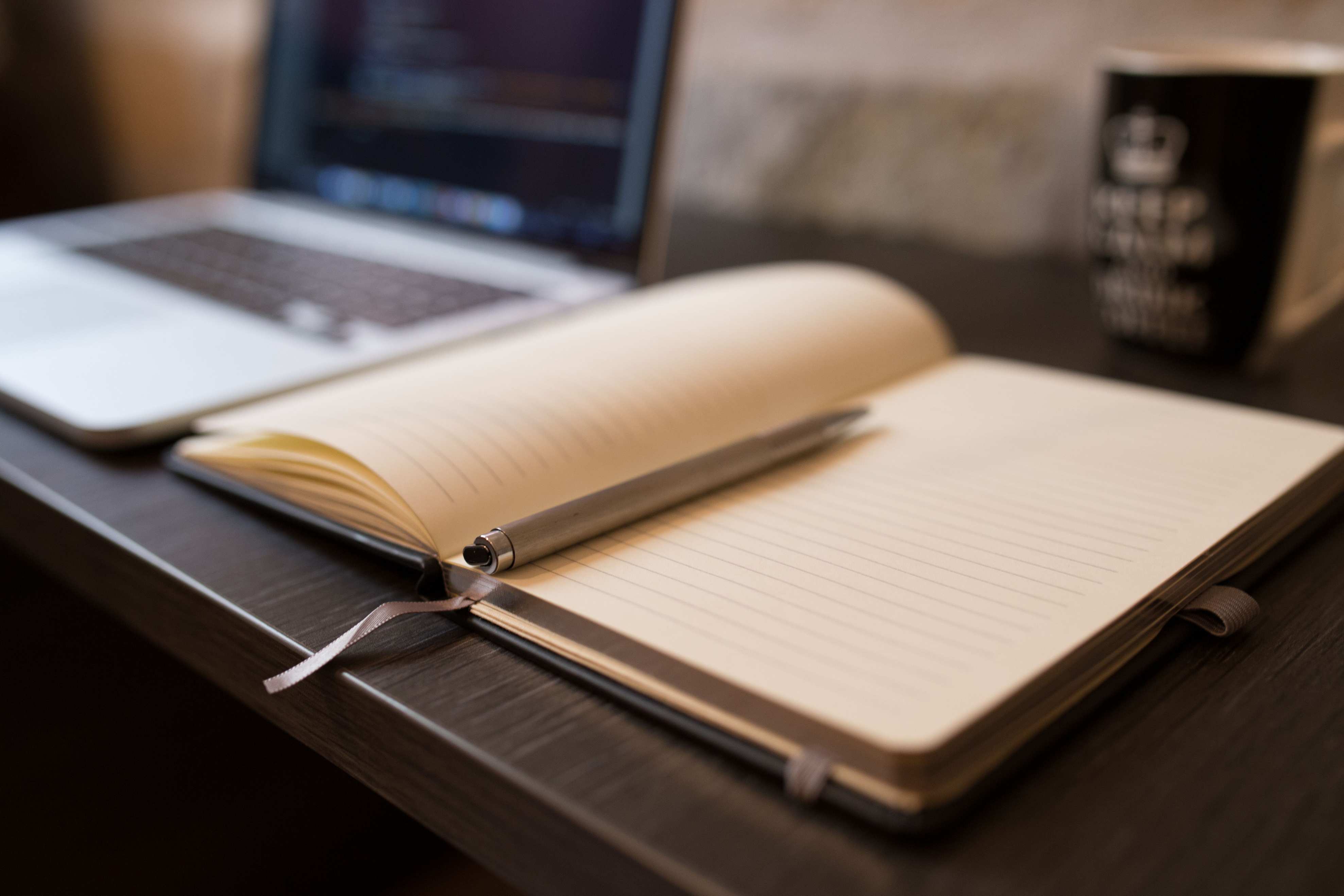 โต๊ะทำงานที่มีแล็ปท็อปในสภาพแวดล้อมห้องทำงาน Open notebook with pen beside a laptop on a wooden desk